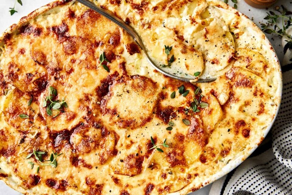 A creamy potato gratin in an oval baking dish, topped with fresh herbs and a metal serving spoon.