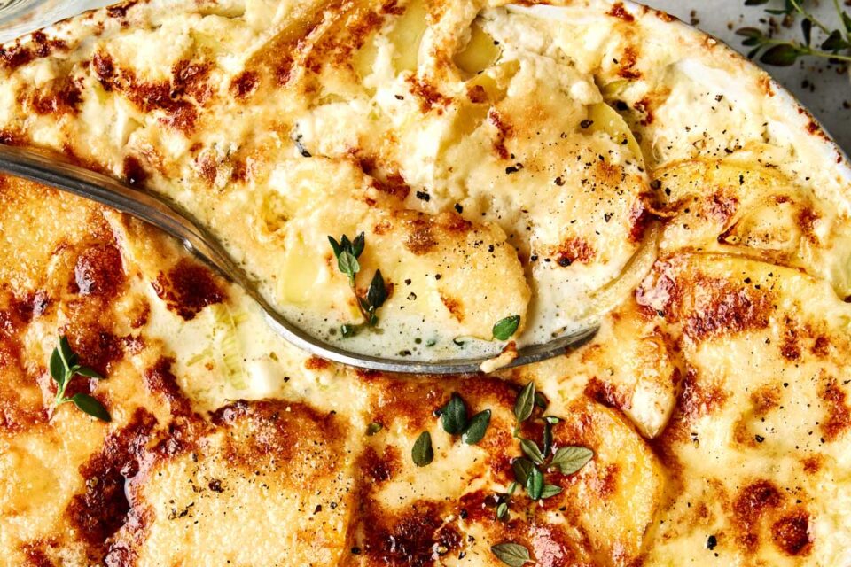 A serving spoon lifting a portion of cheesy au gratin potatoes topped with fresh thyme.