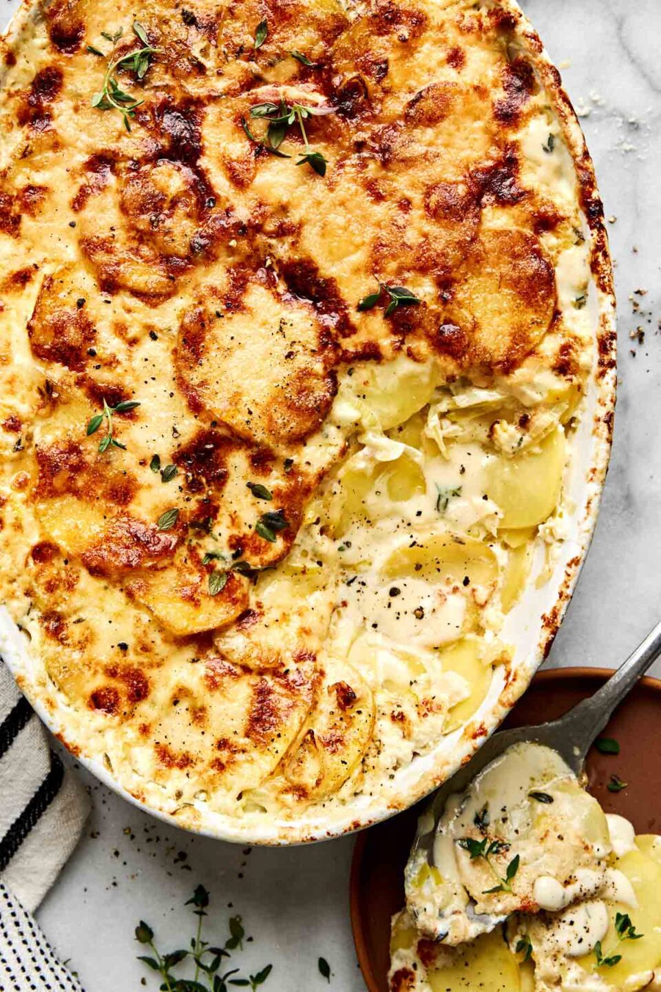 A potatoes ay gratin casserole with a browned, cheesy crust in an oval baking dish, with a serving spoon resting on a nearby plate.