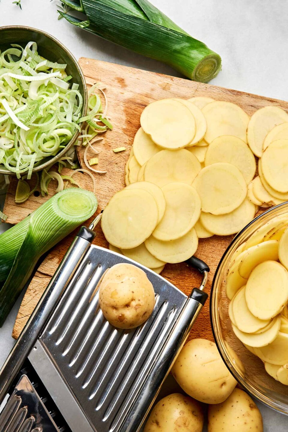 A metal mandoline slicer sits on a wooden cutting board next to a bowl of sliced potatoes, a bowl of sliced leeks, and whole leeks.