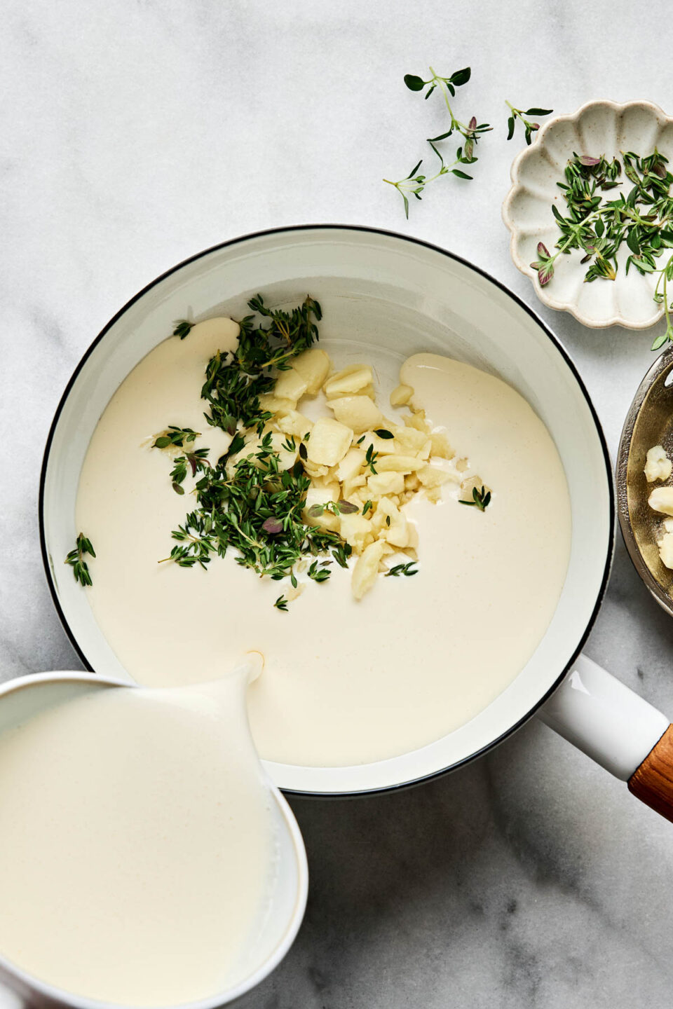 Cream being poured into a saucepan containing chopped garlic and fresh thyme sprigs.