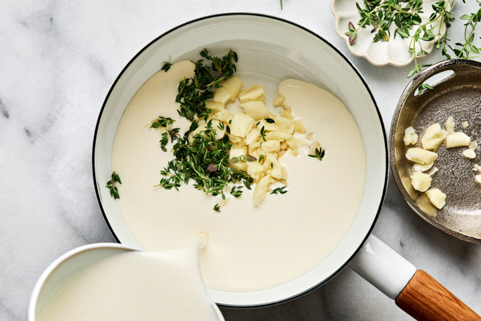 Cream being poured into a saucepan containing chopped garlic and fresh thyme sprigs.