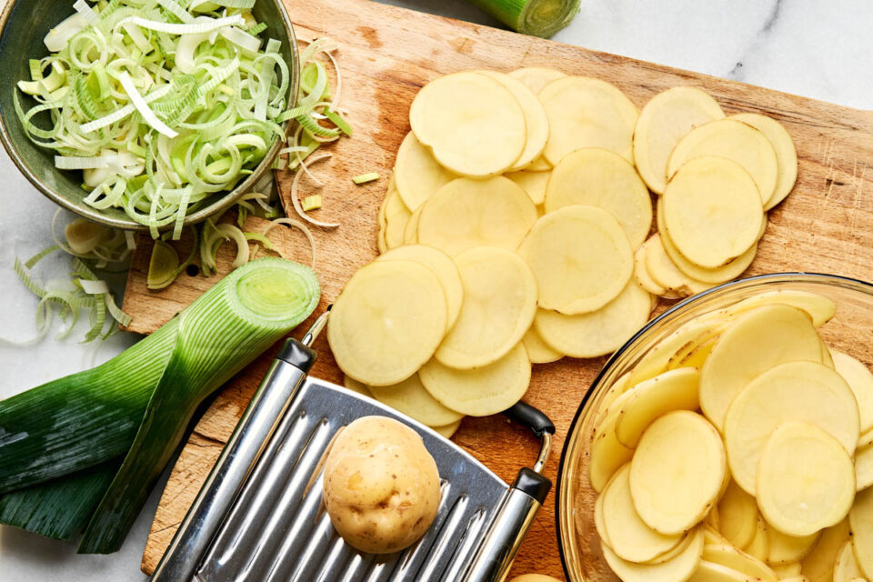 Sliced potatoes and chopped leeks on a wooden cutting board with a mandoline slicer.