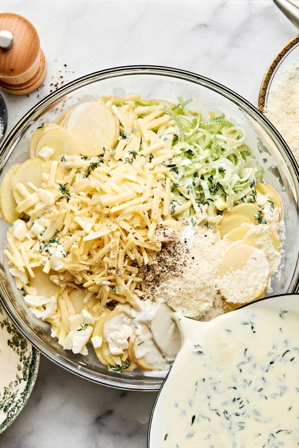A glass bowl filled with sliced potatoes, shredded cheese, and sliced leeks, being poured over with a creamy herb sauce.