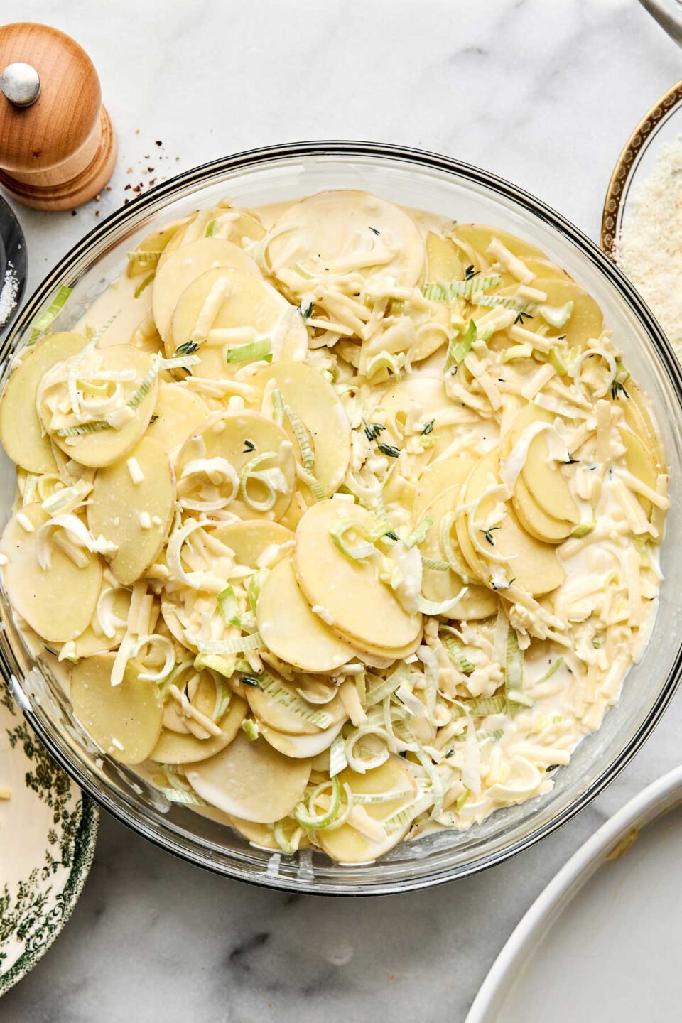 A glass bowl filled with sliced potatoes, shredded cheese, and sliced leeks in a creamy sauce.