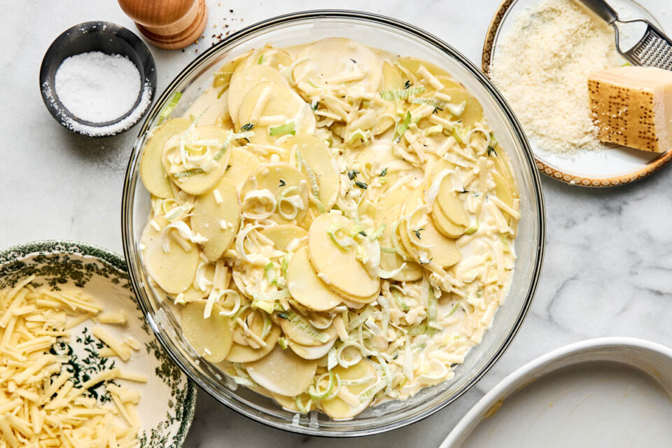 A glass bowl filled with sliced potatoes, leeks, and a creamy sauce, surrounded by small dishes of grated cheese, salt, and pepper.