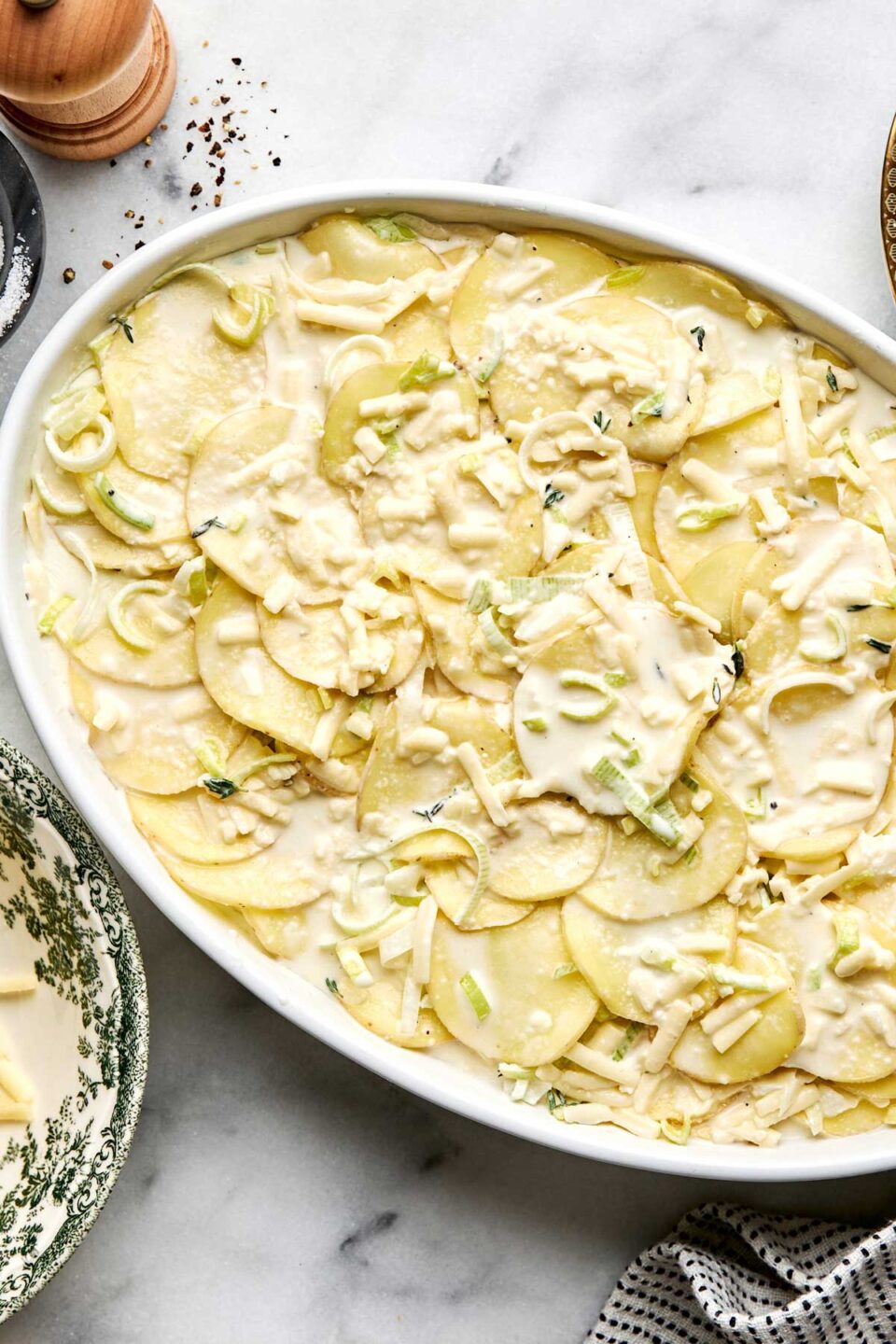 A white baking dish filled with thinly sliced raw potatoes, leeks, and cheese in a creamy sauce, ready to be baked.
