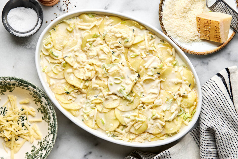An oval baking dish filled with thinly sliced potatoes, leeks, and a creamy sauce, surrounded by bowls of grated cheese, salt, and a piece of Parmesan.